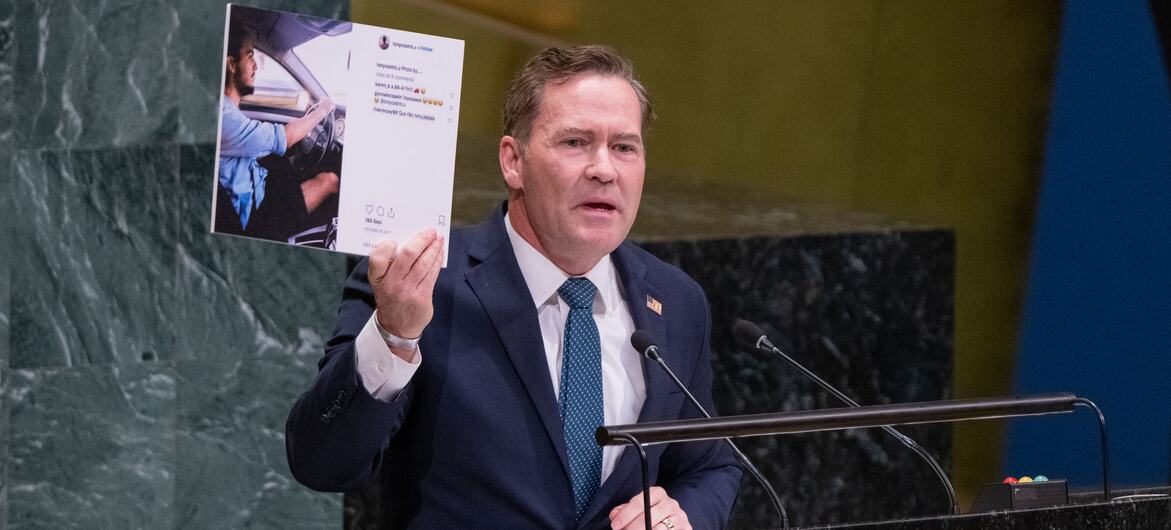 一名身穿深色西装的白人男性外交官在United Nations讲台上发表讲话，同时举着一份打印出来的社交媒体帖子。